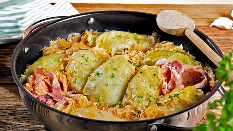 Gefüllte Kohlrouladen mit Speck auf Sauerkraut in einer schwarzen Pfanne, daneben Holzlöffel, Pfeffermühle und Kräuter.