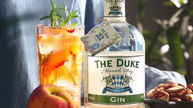 Auf einem Holzbrett stehen eine Gin-Flasche, ein Glas mit Eis und Kräutern, ein Apfel, eine Pfirsichscheibe und eine Schale mit Nüssen.
