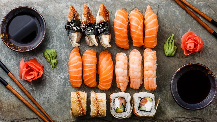 Platte mit Sushi-Variationen, Sojasoßen-Schalen, Wasabi, Ingwerrosen und Essstäbchen auf grauem Untergrund.