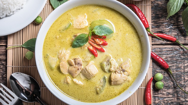 Eine weiße Schale mit grünem Curry, Hühnerfleisch, Basilikum und Chili, daneben ein Teller mit Reis, Limette und rote Chilis.
