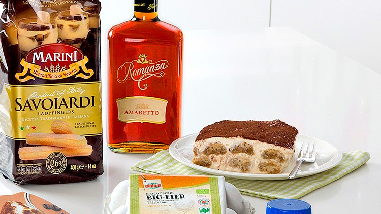 Zutaten für Tiramisu auf weißem Tisch: Löffelbiskuits, Amaretto, Mascarpone, Kakao, Eier, Sahne und fertiges Tiramisu.