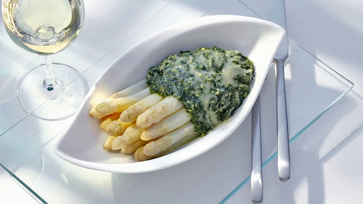 Weißer Spargel mit überbackenem Rahmspinat in einem ovalen Teller, daneben ein gefülltes Weinglas sowie Besteck auf Glasunterlage.