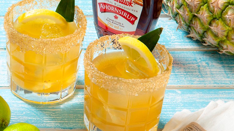 Zwei Cocktails mit Zuckerrand, Zitrone und Ananasblatt vor einer Flasche Ahornsirup, dazu Ananas, Limetten und ein weißes Tuch.