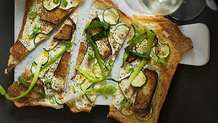 Knuspriger Flammkuchen belegt mit Zucchini, Frühlingszwiebeln, Creme und gebräunten Streifen auf einem Servierbrett.