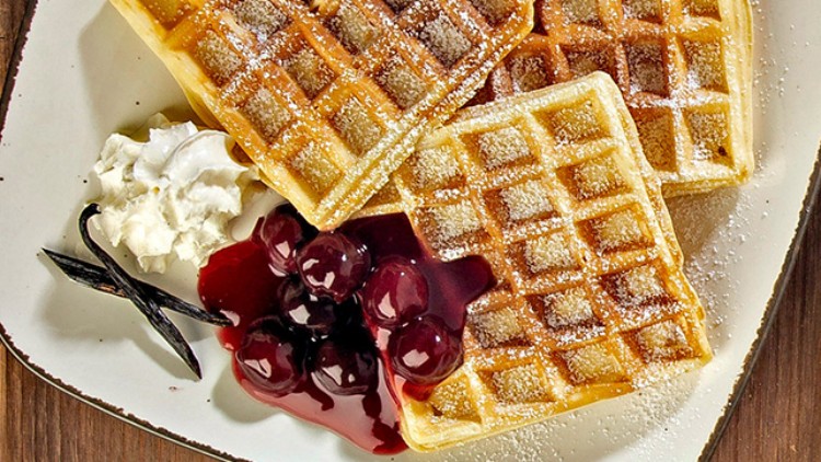 Drei rechteckige Waffeln mit Puderzucker, einer Portion Sahne, einem Vanillestängel und Kirschkompott auf einem Teller.