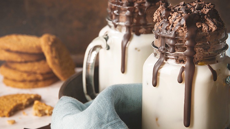 Zwei Gläser mit Milch, Schokoladensauce und Schokostückchen stehen auf einem Tablett, daneben Kekse, Schokolade und eine blaue Stoffserviette.
