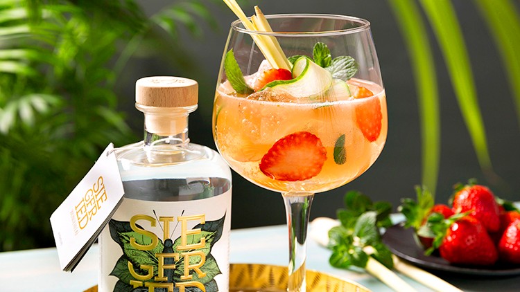 Eine Flasche SIEGFRIED Wonderleaf alkoholfrei und ein gefülltes Weinglas mit Erdbeeren, Gurke und Minze auf einem goldenen Tablett.