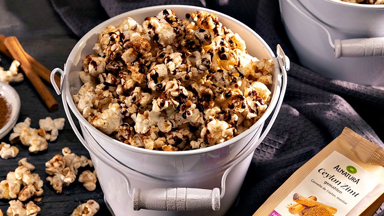 Eimer mit Popcorn, Gewürzpulver in einer weißen Schale, Zimtstangen und eine Packung Alnatura Ceylon-Zimt auf dunklem Untergrund.