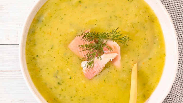 Eine weiße Schale mit grüner Zucchinisuppe, Stück Lachs, Dill und ein gelber Löffel liegt auf einem hellen Holztisch mit Stoffserviette.