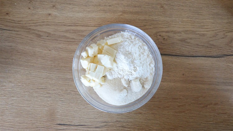Eine Glasschüssel mit Mehl, Zucker und gestückelter Butter steht auf einer Holzoberfläche.