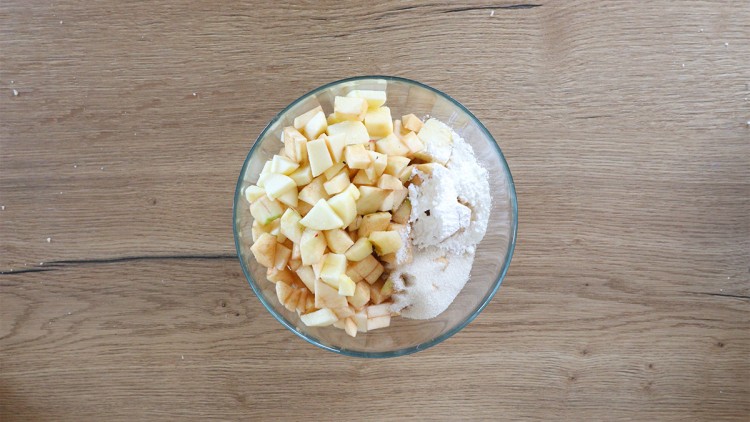 Glas-Schüssel mit klein gewürfelten Äpfeln, Zucker und Mehl auf einem Holztisch.