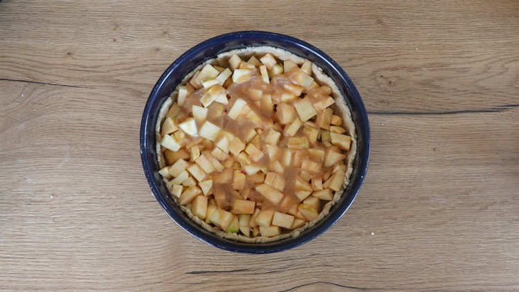 Eine runde Backform mit unbehandeltem Holzrand enthält einen Apfelkuchenrohling mit gewürfelten Äpfeln und heller Sauce.