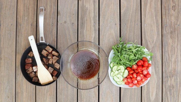 Eine Pfanne mit gebratenen Würfeln, eine Schüssel mit brauner Soße und ein Teller mit Rucola, Tomaten und Gurkenscheiben.