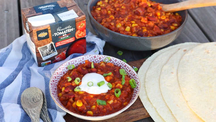 Ein Teller Chili mit Mais und roten Bohnen, garniert mit Frühlingszwiebeln und Crème Fraîche, neben Tortillas und GLOBUS Tomatenpackung.