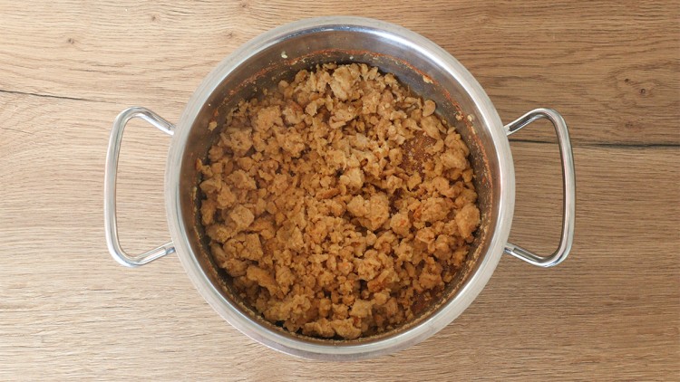 Eine silberne Edelstahlpfanne mit angeröstetem Sojagranulat steht auf einer hellen Holzoberfläche.