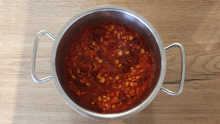 Ein Topf mit rotem Eintopf aus Bohnen, Mais und Tomatensoße steht auf einer Holzoberfläche.