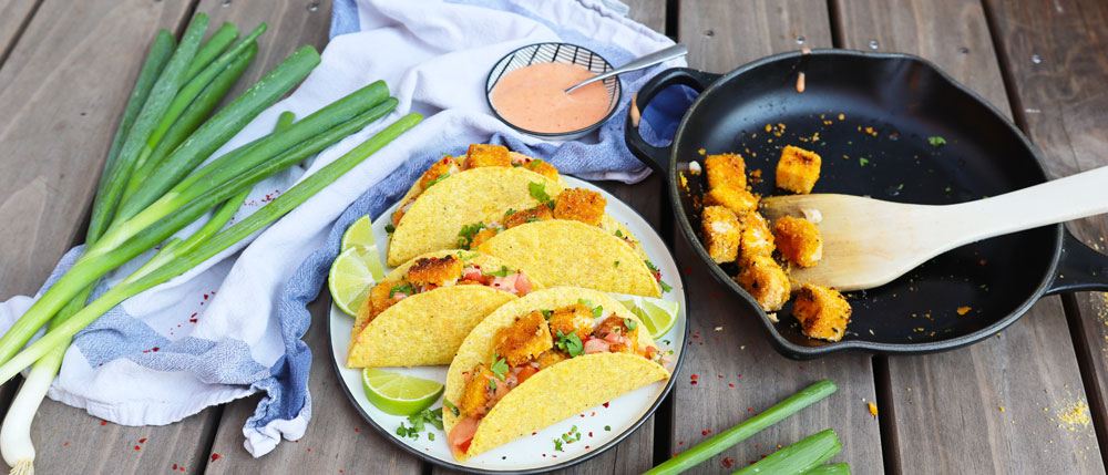 Drei gefüllte Tacos mit Gemüse und gebackenen Würfeln auf Teller, daneben Frühlingszwiebeln, Soße und Pfanne mit Holzlöffel.