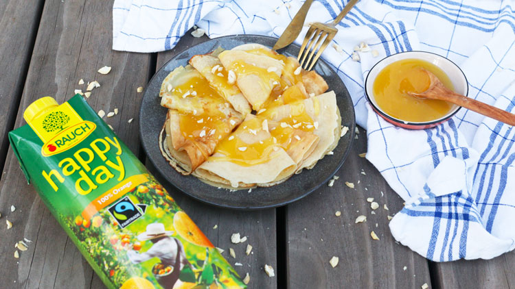 Ein Teller mit Crêpes und gelber Sauce, daneben eine Packung Rauch Happy Day Orangensaft und eine Schale mit Holzlöffel.