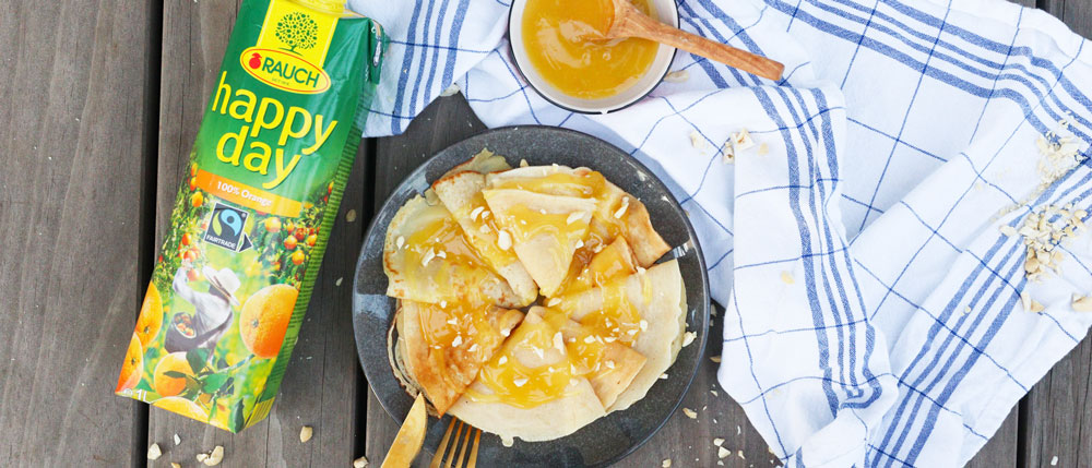 Ein grauer Teller mit Crêpes, Honig und Nüssen, daneben eine Packung Rauch Happy Day Orangensaft und ein Honigglas auf kariertem Tuch.