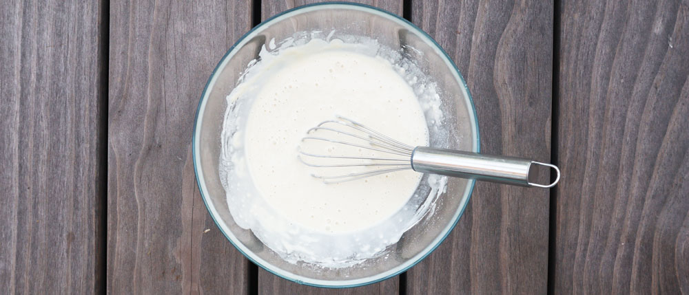 Eine Glasschüssel mit flüssigem, hellen Teig und einem silbernen Schneebesen auf einer dunklen Holzoberfläche.