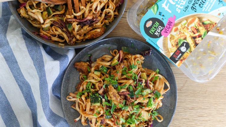 Grauer Teller mit gebratenen Nudeln, Gemüse, veganen Fleischstücken, frischen Kräutern, Verpackung von „Fresh IN Co“ daneben.