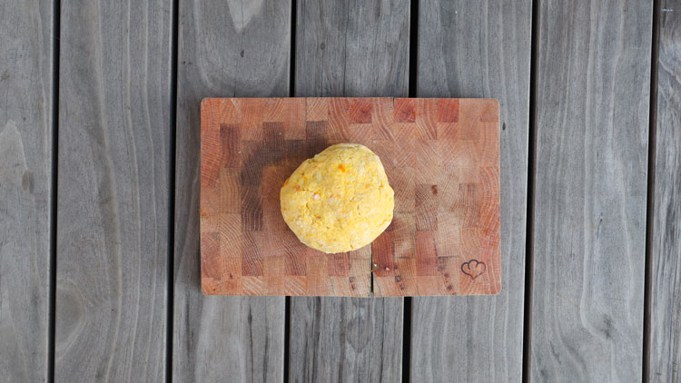 Ein gelber Teigball liegt mittig auf einem Holzschneidebrett mit Herz-Gravur, das auf grauen Holzbrettern steht.