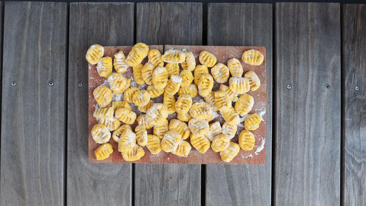 Frische, gelbe Gnocchi liegen auf einem bemehlten Holzbrett, das auf einer Tischplatte aus Holz steht.