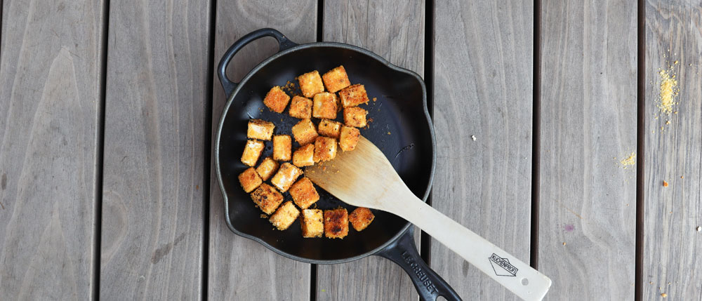 Würfel aus gebratenem Tofu liegen in einer schwarzen Pfanne auf einem Holztisch, ein Holzspatel liegt in der Pfanne.