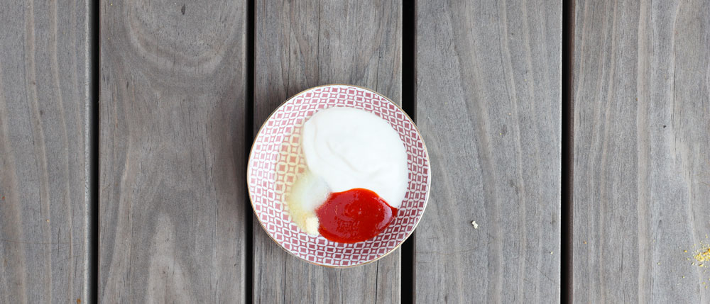Ein kleines Schälchen mit rotem Ketchup, weißem Joghurt und gelbem Senf steht auf einer grauen Holztischplatte.