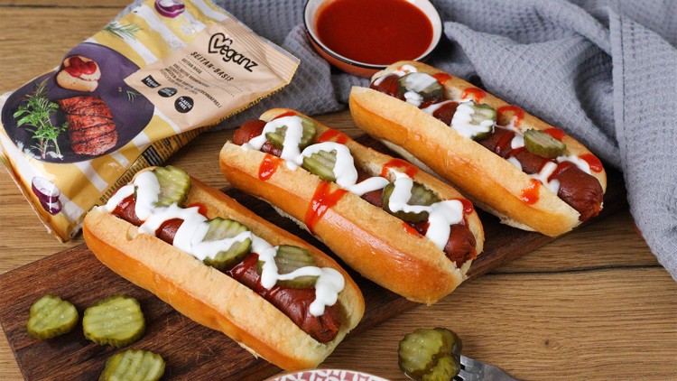 Drei vegane Hotdogs mit Gurkenscheiben, Ketchup und heller Soße auf einem Holzbrett, daneben ein Teller mit Soße und ein Produktbeutel.