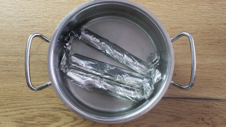Silberner Topf mit zwei Henkeln enthält drei längliche, in Aluminiumfolie eingewickelte Objekte, auf einer Holzoberfläche stehend.
