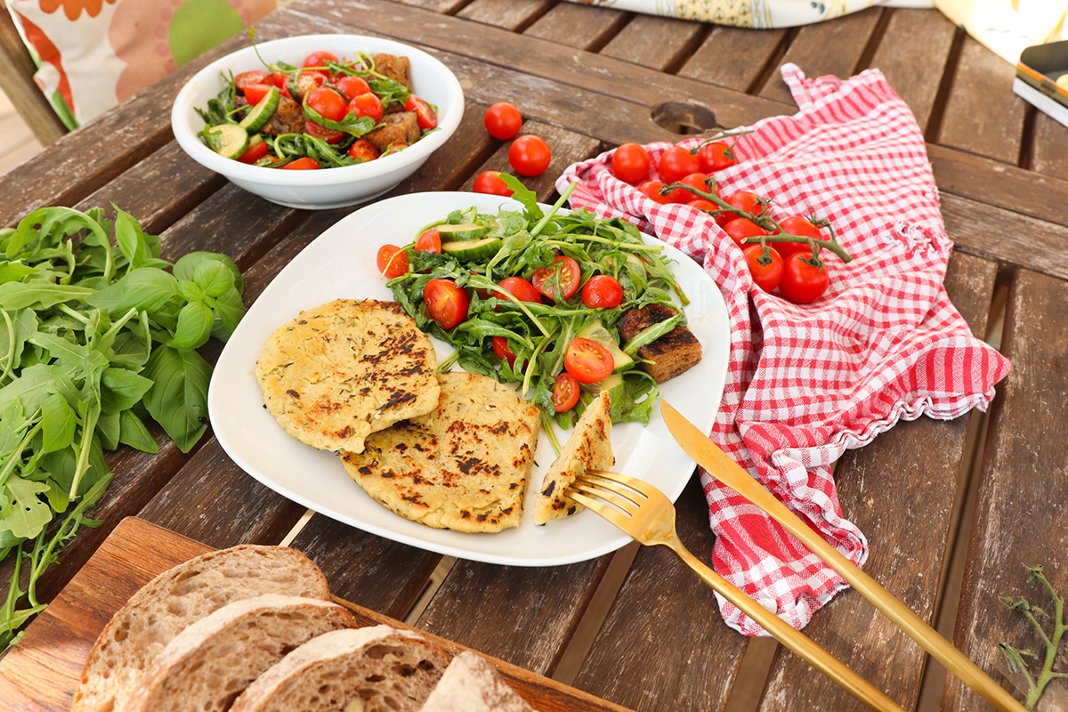 Teller mit Salat aus Rucola, Tomaten und Zucchini, daneben zwei goldbraune Fladen, auf Holztisch mit Brot und Kräutern.