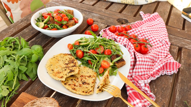 Teller mit Salat aus Rucola, Tomaten und Zucchini, daneben zwei goldbraune Fladen, auf Holztisch mit Brot und Kräutern.