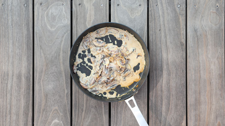 Eine Pfanne mit cremiger Sauce und gebratenen Pilzen steht auf einer hellbraunen Holzoberfläche.