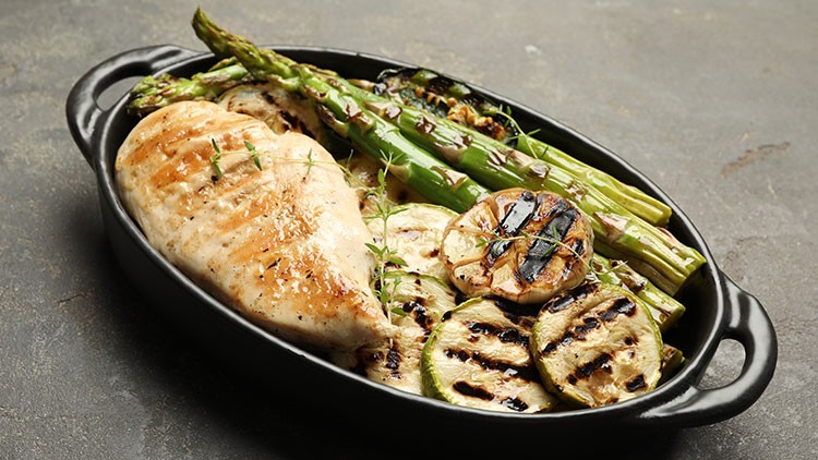 Gegrilltes Hähnchenfilet, Zucchinischeiben, Spargel und eine halbe gegrillte Zitrone in einer schwarzen Auflaufform.