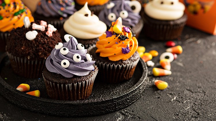 Mehrere Schokoladenmuffins mit buntem Halloween-Dekor wie Augen, Knochen und Zuckerkugeln auf schwarzem Teller.
