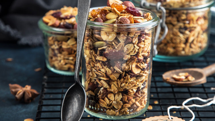 In einem kleinen Glas mit Bügelverschluss liegt knuspriges Müsli mit Nüssen, Kernen und Trockenfrüchten, daneben ein Holzlöffel.