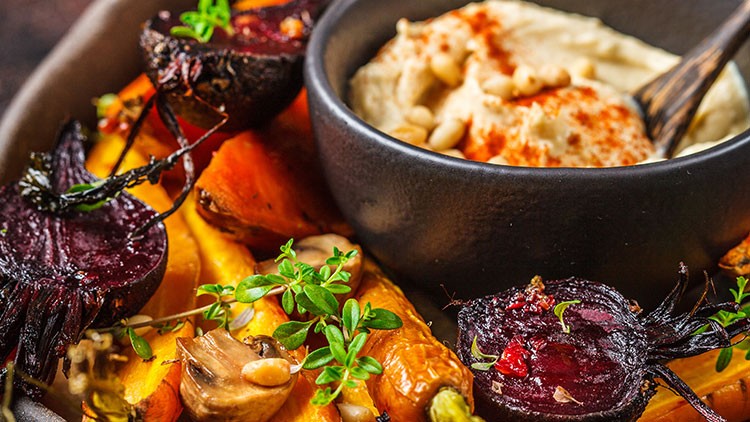 Ein schwarzer Schälchen mit Hummus, dekoriert mit Paprika und Nüssen, steht neben geröstetem Gemüse wie Möhren, Roter Bete und Kräutern.
