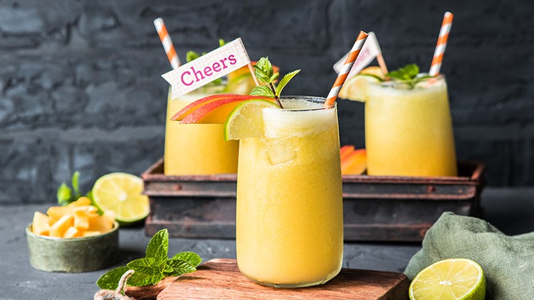 Drei gelbe Smoothies in Gläsern mit gestreiften Strohhalmen, Limettenscheiben und Minze, ein Glas mit Schild „Cheers“.