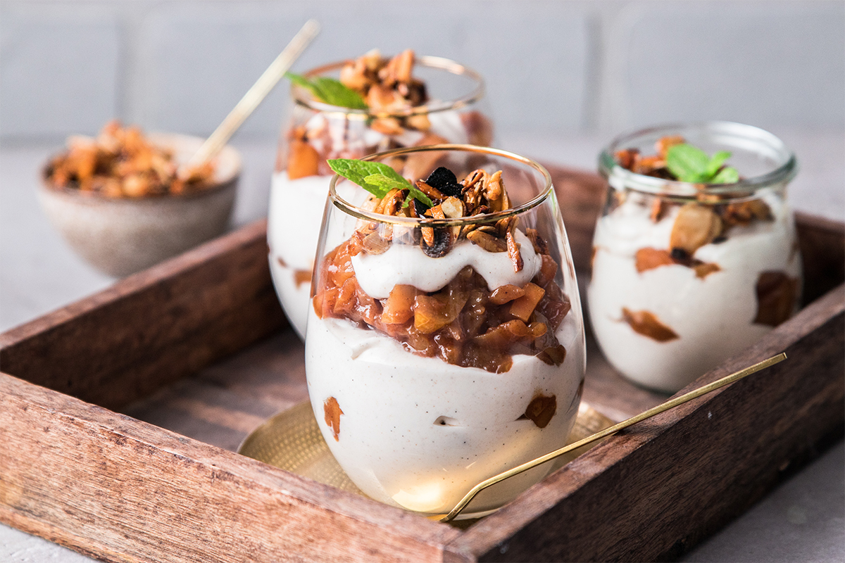 Drei Gläser mit Schichtdessert aus Creme, Apfelstücken, Nüssen und Minzblättern stehen in einem Holztablett.