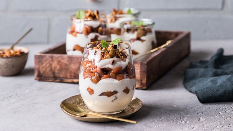 Gläser mit Joghurt, karamellisierten Apfelstücken, Nüssen und Minzblatt auf einem goldenen Teller, im Hintergrund eine Holzschale.
