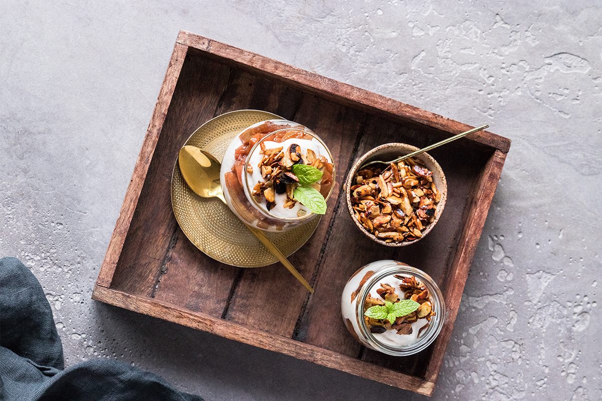 Zwei Dessertgläser mit Creme, gerösteten Nüssen und Minzblättern sowie eine Schale Nüsse auf einem Holztablett mit Löffeln.