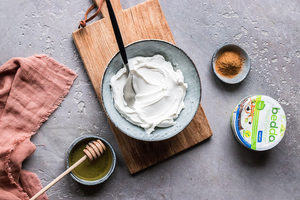 Graue Schale mit weißer Frischcreme, Holzlöffel, Honig, Zimt und „bedda Frischcreme natur“-Packung auf grauem Untergrund.