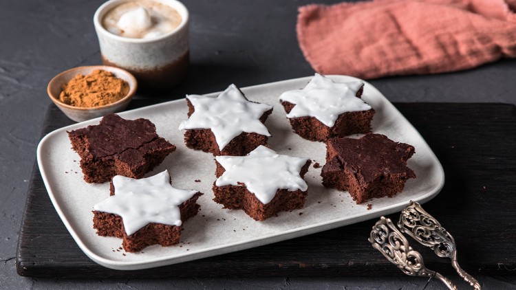 Sechs sternförmige Schokokuchenstücke, teils mit weißem Zuckerguss, auf einer weißen Platte mit dekorativer Zange.