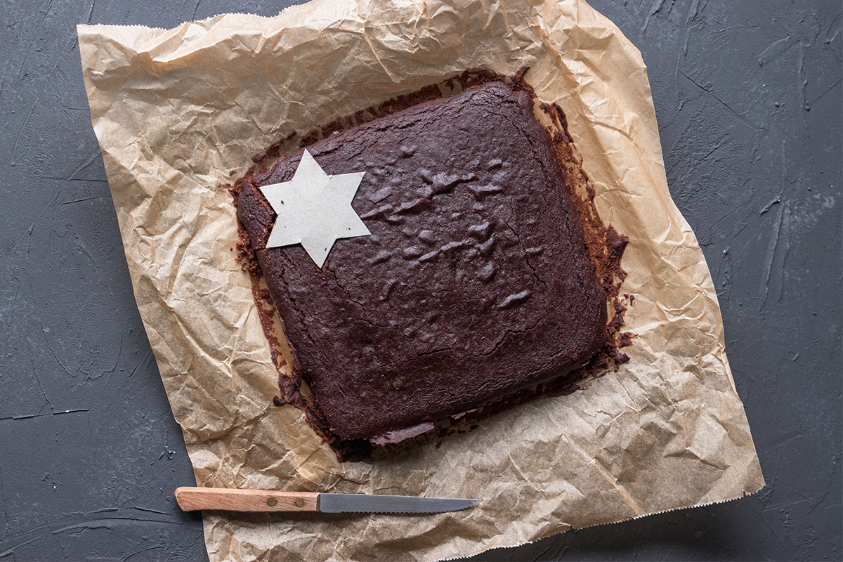 Ein rechteckiger Schokoladenkuchen liegt auf braunem Backpapier, darauf eine weiße Papierstern-Schablone, daneben ein Messer.