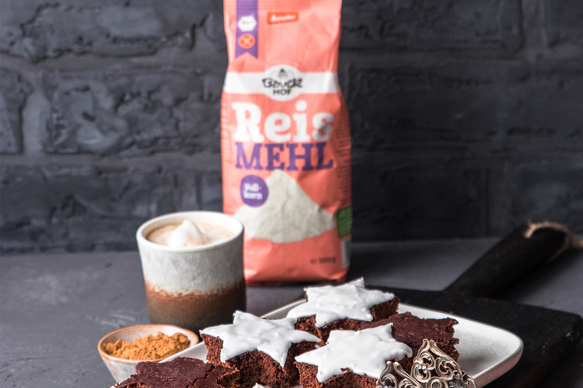 Eine Packung Bauckhof Reismehl, ein Keramikbecher mit Schaum, Zimt und sternförmige Lebkuchen auf einem Teller.