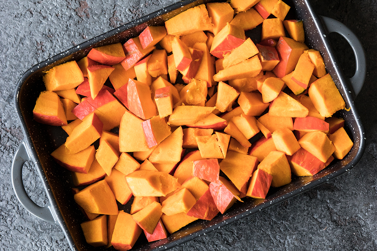 Würfel aus orangefarbenem Kürbis mit Schale liegen in einer schwarzen, rechteckigen Auflaufform mit silbernen Griffen.