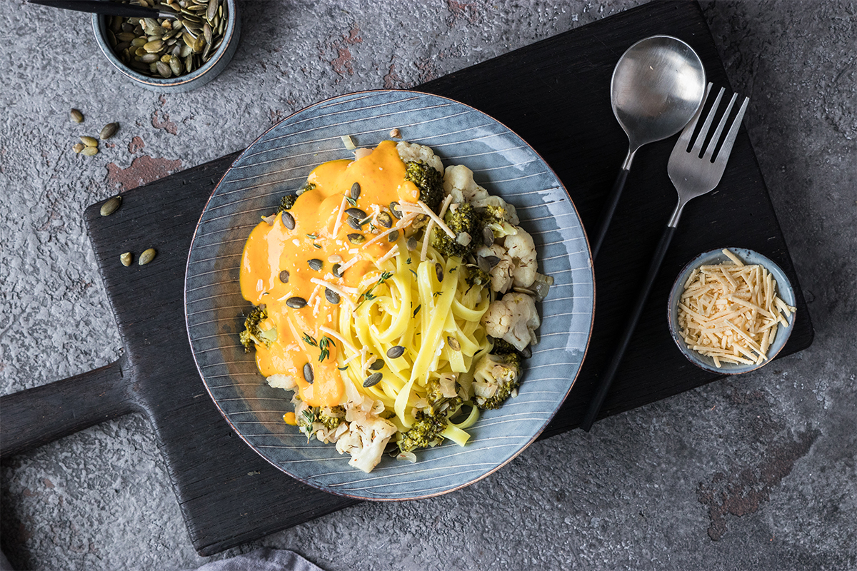 Blauer Teller mit Tagliatelle, Brokkoli, Blumenkohl, Käse-Soße und Kürbiskernen auf schwarzem Brett, daneben Besteck und Käse.