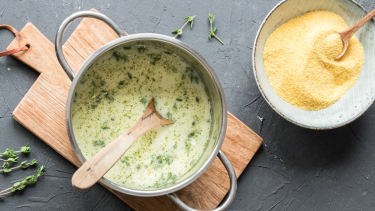 Topf mit cremiger Suppe und grünem Gemüse auf Holzbrett, daneben eine Schüssel mit gelbem Maisgrieß und Holzlöffel.