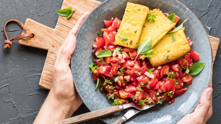 Ein Teller mit frischem Tomatensalat und drei gebratenen Polentascheiben wird von zwei Händen über einem Holzbrett gehalten.